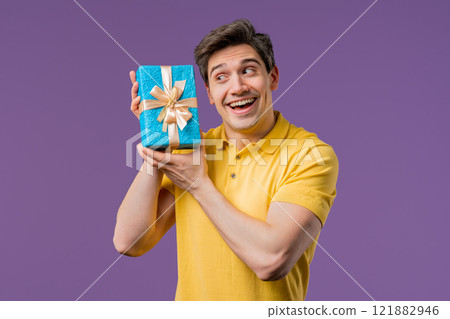 Handsome man holding gift box on violet studio background and smiles to camera. Happy european guy received present and interested in what's inside. 121882946