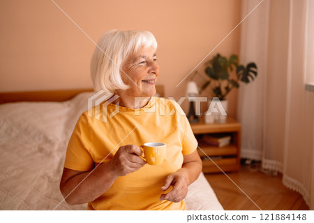 Caucasian blonde senior woman sitting on a white bed with a cup in her hand. Female relaxing and drinking cup of hot coffee or tea in the bedroom. Caucasian blonde senior woman sitting on a white bed with a cup in her hand. Female relaxing and drinking cup of hot coffee or tea in the bedroom. 121884148