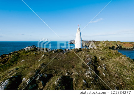 Coastal scene. A white, conical landmark stands on a rocky outcrop overlooking a calm sea and distant coastline under a clear blue sky. A small red marker tops the structure. Coastal scene. A white, conical landmark stands on a rocky outcrop overlooking a calm sea and distant coastline under a clear blue sky. A small red marker tops the structure. 121884277