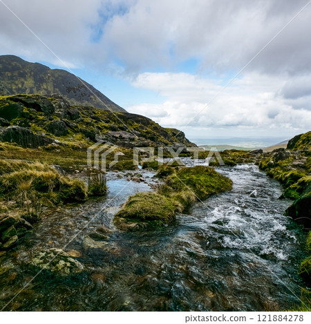 A fast-flowing stream navigates a rocky, grassy mountain pass. The sky is partly cloudy. 121884278