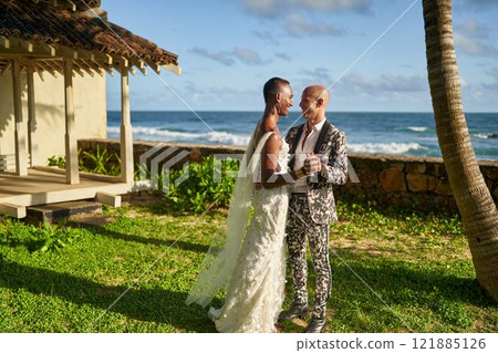 LGBTQ couple in wedding clothes by tropical villa. Ambiguous gender person in white dress as bride holds hands with groom in floral suit. Beach ceremony vibe. Same-sex wedding concept. 121885126