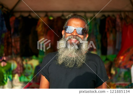 Senior artist with grey beard smiles at craft fair. Wears tie-dye t-shirt. Sunglasses reflect vibrant booths. Creative stall display. Daytime outdoor event. Senior artist with grey beard smiles at craft fair. Wears tie-dye t-shirt. Sunglasses reflect vibrant booths. Creative stall display. Daytime outdoor event. 121885134