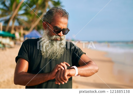 Senior stylish man with gray beard, sunglasses checks fitness tracker on sunny beach. Modern elder enjoys tech gadgets, stays active outdoors. Trendy, healthy lifestyle choice highlights independence. 121885138