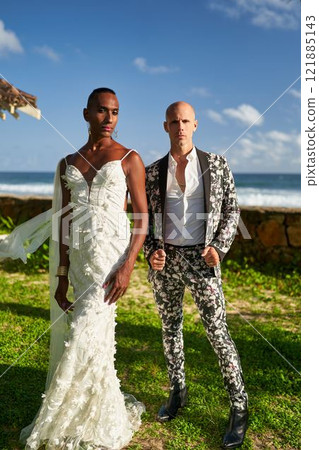 Ambiguous gender fluid ethnic black person in white dress with partner in floral suit at tropical villa wedding. LGBTQ couple celebrates pride, love by sea. Bold, diverse representation. 121885143