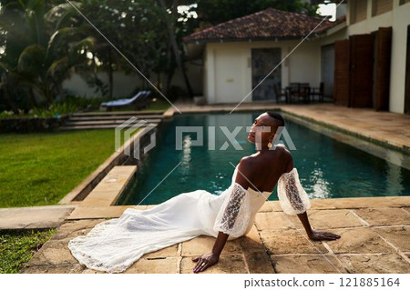 Black person in white bridal dress sits by villa pool. Gender fluid bride at luxury outdoor wedding venue. LGBTQ wedding fashion and serenity. Modern inclusive ceremony. Black person in white bridal dress sits by villa pool. Gender fluid bride at luxury outdoor wedding venue. LGBTQ wedding fashion and serenity. Modern inclusive ceremony. 121885164