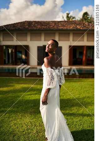 Smiling person in white wedding dress walks on villa lawn under blue sky. Gender fluid black bride celebrates LGBTQ wedding. Summer outdoor ceremony. Elegant bridal fashion. 121885168