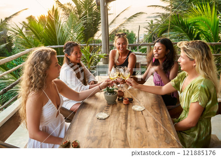 Diverse women toast, enjoy friendship at seaside restaurant. Sunset view. Girls celebrate event. Warm atmosphere, coastal vibes. Relaxed summer dining. Perfect for lifestyle, travel, hospitality. Diverse women toast, enjoy friendship at seaside restaurant. Sunset view. Girls celebrate event. Warm atmosphere, coastal vibes. Relaxed summer dining. Perfect for lifestyle, travel, hospitality. 121885176