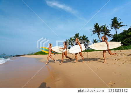 Friends walk on tropical beach holding surfboards. Young surfers explore ocean waves under blue sky. Adventure surf camp. Summer travel, beach sports. Female surfers enjoy sun, sand in paradise. Friends walk on tropical beach holding surfboards. Young surfers explore ocean waves under blue sky. Adventure surf camp. Summer travel, beach sports. Female surfers enjoy sun, sand in paradise. 121885182