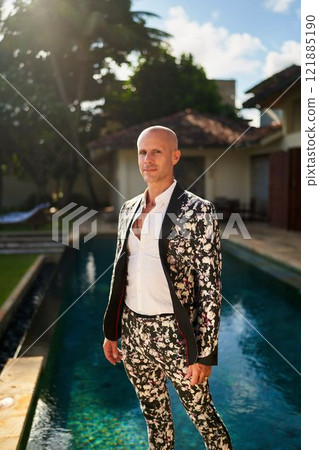 Bald man in bold floral suit stands at villa poolside. Daytime event, stylish attire with open shirt. Fashion trend. Possible gay groom outfit. Elegant garden setting. 121885190