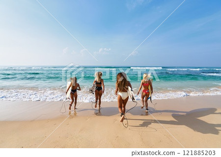Four women run to ocean with surfboards on sandy beach. Group of friends enjoy sunny day at sea, ready for surfing, embracing active lifestyle. Summer vacation outdoors, water sports enthusiasts. 121885203