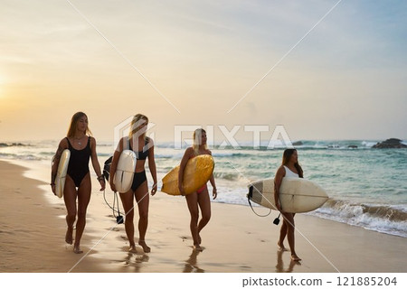 Four women walk on beach holding surfboards. Sunset sky, ocean waves, golden sand. Female surfers enjoy seaside, summer sport and leisure. Group of friends, fit lifestyle, coastal adventure. 121885204