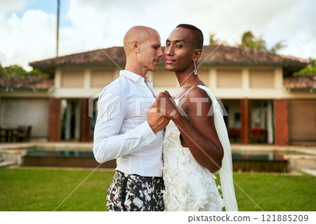 LGBTQ wedding at tropical villa. Ambiguous gender fluid bride in white dress, groom in bold suit dance. Elegant couple in vibrant love story. Inclusive, diverse celebration. Modern wedding. 121885209