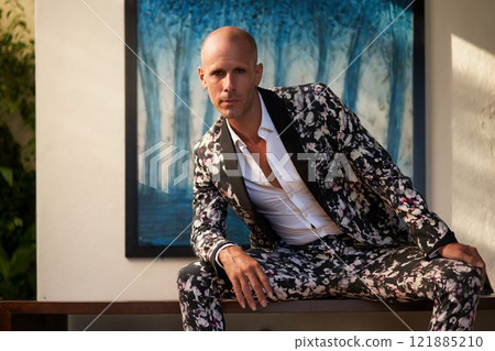 Bald man in floral suit sits confidently at luxury villa. Bold fashion wedding style, groom attire concept. Sunlit elegant setting. Sophisticated outfit trend for modern nuptials. 121885210