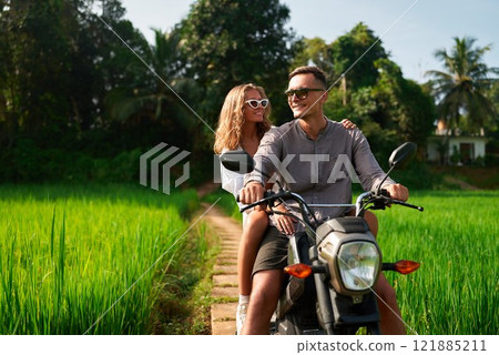 Couple rides motorbike through green rice field path. Man drives, woman sits behind enjoying tropical scenery. Smiling duo explores exotic travel destination on sunny day. Adventure seekers in nature. 121885211