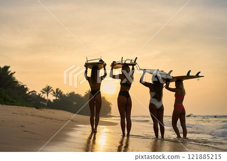 Four surfers walk along beach holding surfboards at sunset. Women enjoy ocean, waves, tropical adventure. Friends explore travel, prepare for surfing. Relax, nature, sea, sky inspire vacation. 121885215