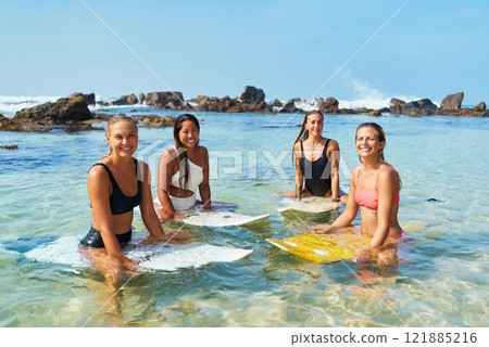 Group of diverse young women in swimsuits with surfboards relax in clear ocean waters near rocky shore under blue sky. Friends enjoy sunny beach day. Summer leisure activity. 121885216