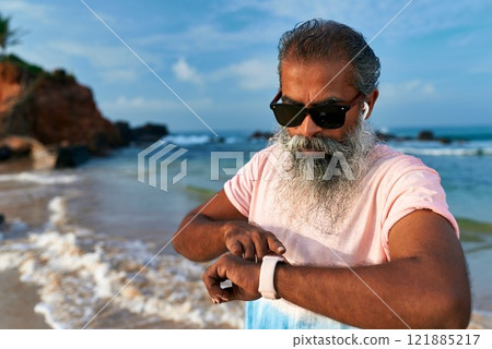 Stylish senior with gray beard at beach uses smart watch and wireless earbuds. Elderly man enjoys modern tech. Relaxed grandpa embraces fitness gadget lifestyle. Stylish senior with gray beard at beach uses smart watch and wireless earbuds. Elderly man enjoys modern tech. Relaxed grandpa embraces fitness gadget lifestyle. 121885217