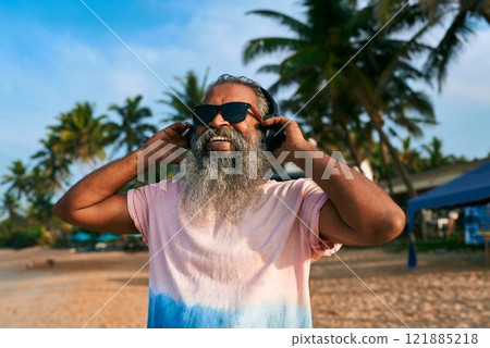 Senior ethnic man with gray beard enjoys music at sunny tropical beach wearing sunglasses and headphones. Stylish elderly dancing, uplifting mood, sunny leisure. Summer joy vibes, retro style cool. Senior ethnic man with gray beard enjoys music at sunny tropical beach wearing sunglasses and headphones. Stylish elderly dancing, uplifting mood, sunny leisure. Summer joy vibes, retro style cool. 121885218