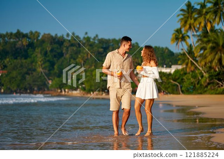 Romantic couple walks on sandy beach with drinks, enjoying sunset. Man and woman on honeymoon stroll by sea, tropical getaway. Love, vacation, dating app match, happiness in serene seaside setting. Romantic couple walks on sandy beach with drinks, enjoying sunset. Man and woman on honeymoon stroll by sea, tropical getaway. Love, vacation, dating app match, happiness in serene seaside setting. 121885224