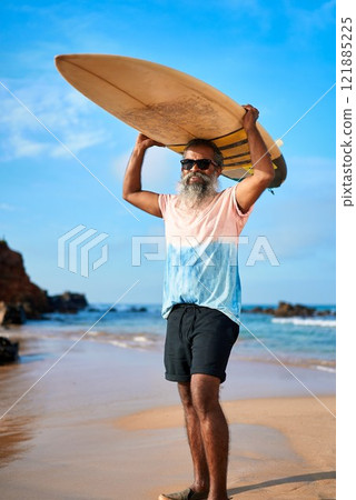 Active senior man holds surfboard on sunny beach. Ethnic male in stylish outfit, surfing. Older surfer enjoys adventure, smiles at sea. Healthy lifestyle at coast. Image for travel sites. Active senior man holds surfboard on sunny beach. Ethnic male in stylish outfit, surfing. Older surfer enjoys adventure, smiles at sea. Healthy lifestyle at coast. Image for travel sites. 121885225