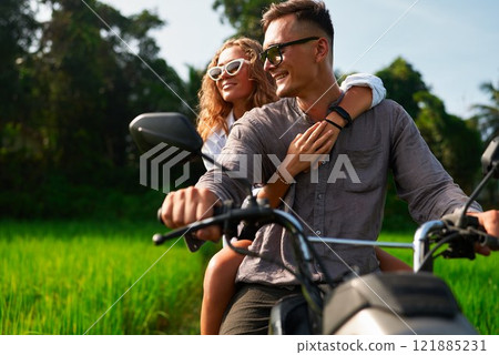 Happy couple ride motorbike through lush rice fields. Woman hugs man from behind. Romantic adventure in nature. Engagement or honeymoon trip. Celebrate love on motorcycle journey. Happy couple ride motorbike through lush rice fields. Woman hugs man from behind. Romantic adventure in nature. Engagement or honeymoon trip. Celebrate love on motorcycle journey. 121885231