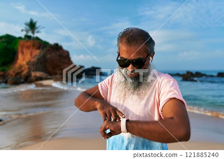 Senior Sri Lankan man with fitness tracker stands on beach. Uses smartwatch for training, wellbeing, wearing wireless earbuds. Modern tech enhances health. Ocean view, rocky shore. Healthy lifestyle. 121885240