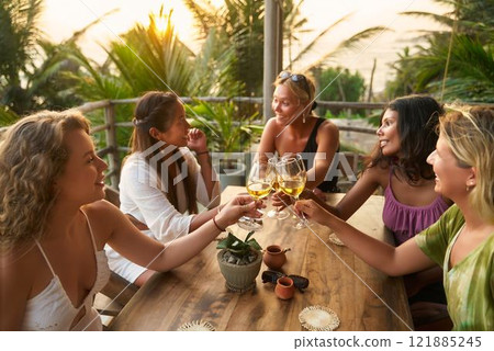 Diverse women at tropical restaurant, toast with wine glasses, relax on girls trip. Celebrate bachelorette party, enjoy sunset moment, happy occasion with friends in exotic setting. 121885245
