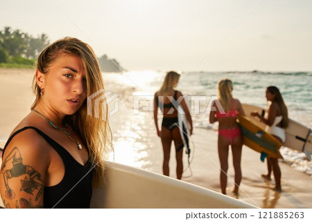 Young women surfers on sandy beach with surfboards. Tattoos, ocean waves, sunset in background. Adventurous friends chat, ready to catch waves. Summer beach vibes, surfing lifestyle. Young women surfers on sandy beach with surfboards. Tattoos, ocean waves, sunset in background. Adventurous friends chat, ready to catch waves. Summer beach vibes, surfing lifestyle. 121885263