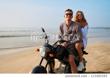 Young couple on motorbike by sea beach. Man with sunglasses drives motorcycle along shore. Woman sits behind, enjoying ocean views. Romantic travel adventure on island. Perfect for vacation, filming. 121885265