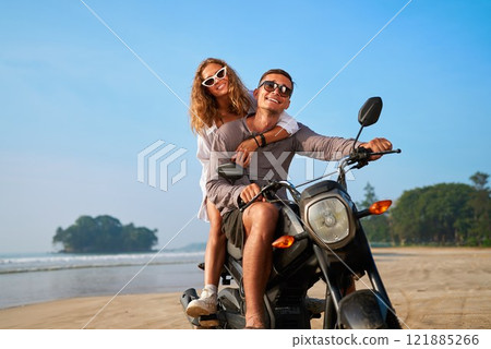 Couple rides motorbike on beach. Man drives motorcycle, woman hugs behind. Casual clothes, sunglasses. Relaxing tropical coast vacation, nature adventure. Freedom, leisure time. 121885266
