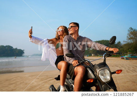 Couple on tropical beach ride scooter. Woman in bikini, guy in casual shirt. Take selfie, smile on sandy ocean shore. Travel influencers explore exotic place. Adventure, freedom mood. 121885267