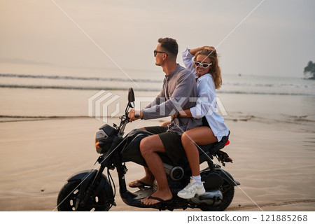 Young couple on motorbike at beach shore. Romantic travel, adventure on motorcycle during sunset. Lovers enjoy ocean freedom. Happy vacation vibes. Rider drives scooter, girlfriend holds, smiles. 121885268