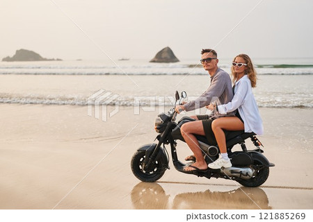 Couple rides scooter by ocean shore. Man drives, woman sits behind in sunglasses. They explore beach on motorbike during sunset. Relaxing budget travel adventure. 121885269