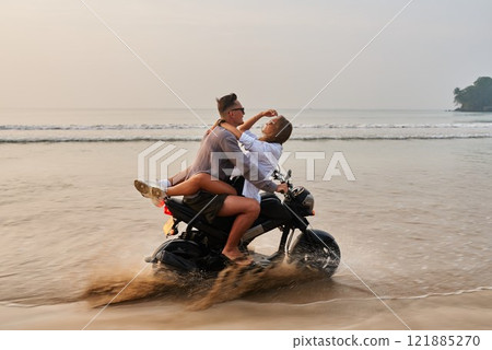 Couple rides motorbike on sandy beach. Man drives motorcycle while woman spreads arms. Lovers enjoy romantic beach vacation at sunset. Adventure and freedom vibes. 121885270