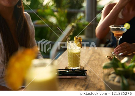 Two women enjoy drinks at outdoor cafe. Pineapple cocktail and margarita on wooden table. Relax in tropical setting with lush plants. Social dining and summer vibes. 121885276