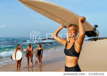Smiling woman with surfboard on sandy ocean beach. Group of surfers in swimwear discuss waves. Summer surfing camp. Ocean sports, adventure travel, beach vibes. 121885282