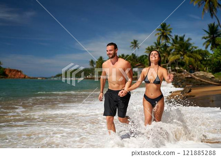Fit couple runs through surf on tropical beach. Man and woman in swimwear enjoy seaside run, sunny adventure. Healthy joggers splash water, exercise during vacation. Active lifestyle, ocean jog. 121885288
