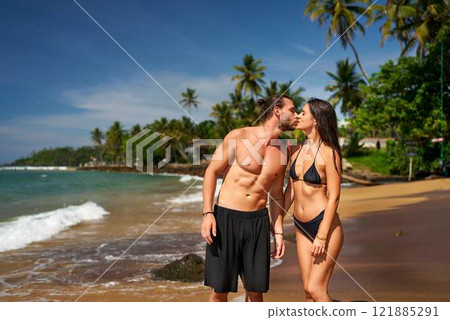Romantic couple kissing on tropical beach, man and woman in swimwear enjoying honeymoon by sea. Fit pair with toned bodies sharing tender moment under palm trees, love on sunny coastline. Romantic couple kissing on tropical beach, man and woman in swimwear enjoying honeymoon by sea. Fit pair with toned bodies sharing tender moment under palm trees, love on sunny coastline. 121885291