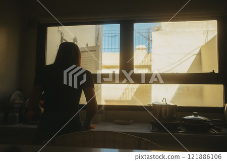 A woman is gazing out of a window located in a kitchen area A woman is gazing out of a window located in a kitchen area 121886106