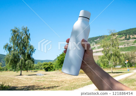 Man holding in the hand white thermo stainless bottle with refreshing water on the nature. Man holding in the hand white thermo stainless bottle with refreshing water on the nature. 121886454