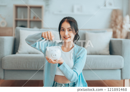Young Asian woman holding piggy bank and sitting on the floor at home 121891468