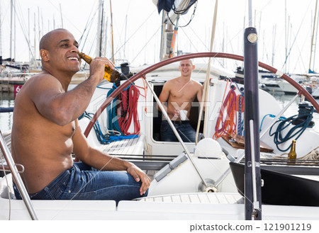 Man topless with a bottle of beer in his hand sits on the deck of a yacht in seaport 121901219