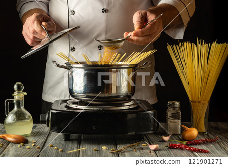 A passionate chef adds spices to boiling water in a pot of spaghetti. Aromatic ingredients surround him, creating a warm atmosphere. The kitchen shines with culinary skill A passionate chef adds spices to boiling water in a pot of spaghetti. Aromatic ingredients surround him, creating a warm atmosphere. The kitchen shines with culinary skill 121901271