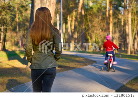 Mother in green jacket watches child riding bicycle 121901512