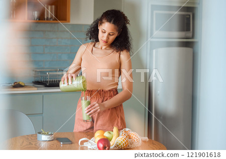 Healthy, nutrition and wellness smoothie made by young woman for her fresh, green detox or vegan diet at home in her kitchen. Vegetarian female pouring and drinking her organic homemade fruit juice 121901618