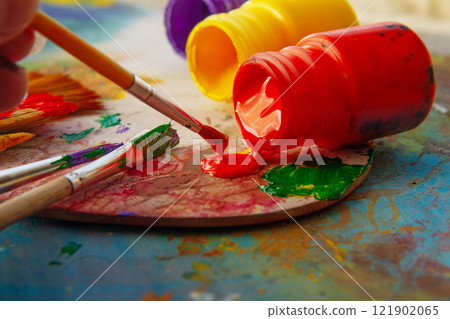 Tip of water color brush is dipped in red paint on palette among lying paint cans 121902065