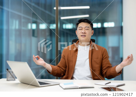 Mature Asian man resting meditating sitting in lotus position inside office. Businessman working with laptop at workplace, doing breathing exercises. Mature Asian man resting meditating sitting in lotus position inside office. Businessman working with laptop at workplace, doing breathing exercises. 121902417