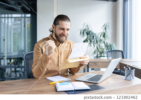 A young professional in a modern workspace joyfully reacts to positive news, expressing delight, motivation, and achievement. The setup bridges productivity with optimism. A young professional in a modern workspace joyfully reacts to positive news, expressing delight, motivation, and achievement. The setup bridges productivity with optimism. 121902462