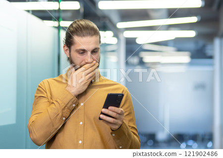 A man wearing a mustard shirt looks at a smartphone with concern in an office environment. A man wearing a mustard shirt looks at a smartphone with concern in an office environment. 121902486