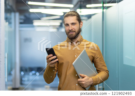 Young professional dressed in business casual attire holds a laptop and smartphone while standing in a modern office environment, showcasing confidence and tech-savviness. Young professional dressed in business casual attire holds a laptop and smartphone while standing in a modern office environment, showcasing confidence and tech-savviness. 121902493